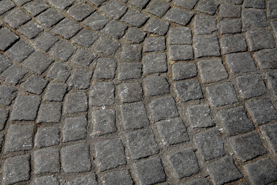 Close-up view of a cobbled street surface with irregularly shaped, flat stone paving slabs in shades of gray and beige, arranged in a random pattern, with narrow gaps filled with dark mortar. The ston