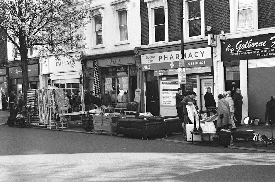 Two removal workers are engaged in the loading process outside a shop on Bermondsey high street, where a large delivery truck with an open rear door is parked on the pavement. The workers are handling