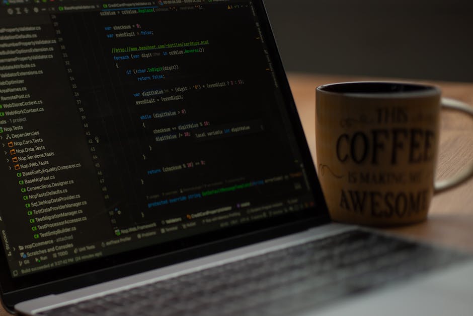 A person typing on a black Dell laptop placed on a wooden desk, with the screen displaying lines of code in a programming environment. To the left of the laptop, there is a black DSLR camera with a le