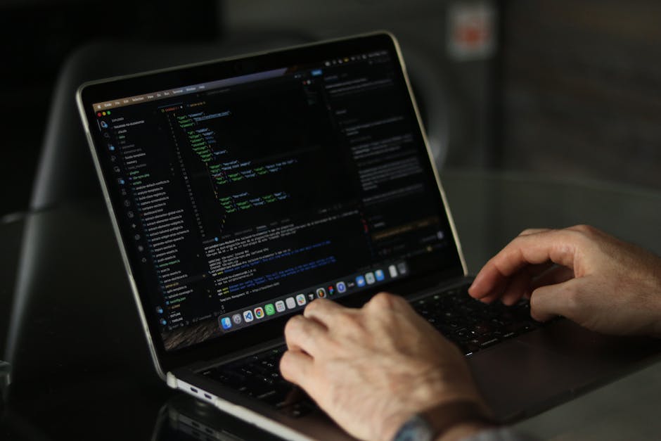 A close-up image of a person's hand operating a laptop computer on a wooden surface. The laptop screen displays lines of code in a text editor, indicating programming or technical work. The individual