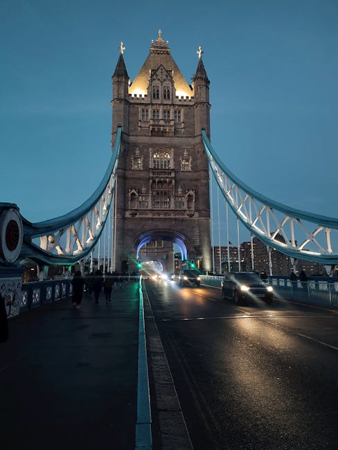 A long exposure photograph of Tower Bridge in Bermondsey during twilight shows the bridge's iconic Gothic-style towers with illuminated windows and decorative turrets, set against a deep blue evening 