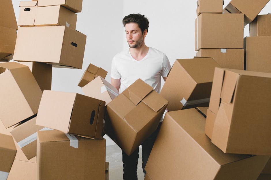 A man wearing a white t-shirt and blue jeans is standing inside a room filled with numerous cardboard moving boxes of various sizes, some sealed with packing tape and others open with packing material